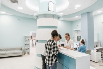 Patients stand at the front desk of a white, sterile office as a member of the front desk staff assists them.